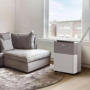Air purifier next to a gray sectional sofa in a room with large windows.