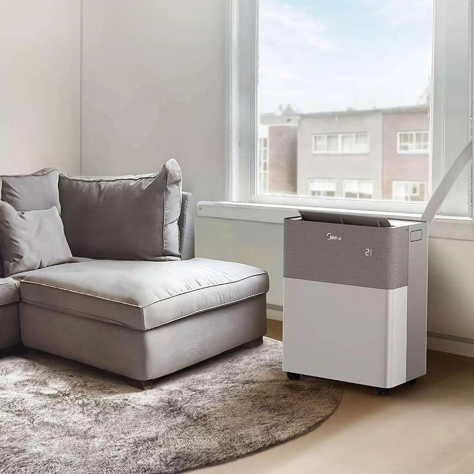 Air purifier next to a gray sectional sofa in a room with large windows.